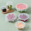 Clear silicone stretch lids covering a watermelon half, glass bowls of fruit, a jar and a glass on a sage green table