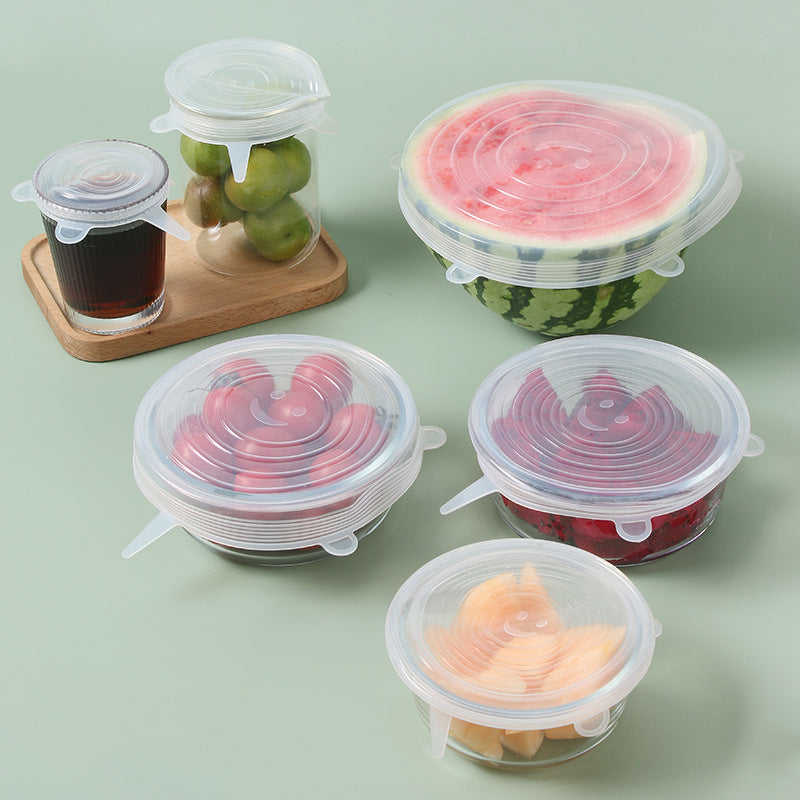 Clear silicone stretch lids covering a watermelon half, glass bowls of fruit, a jar and a glass on a sage green table