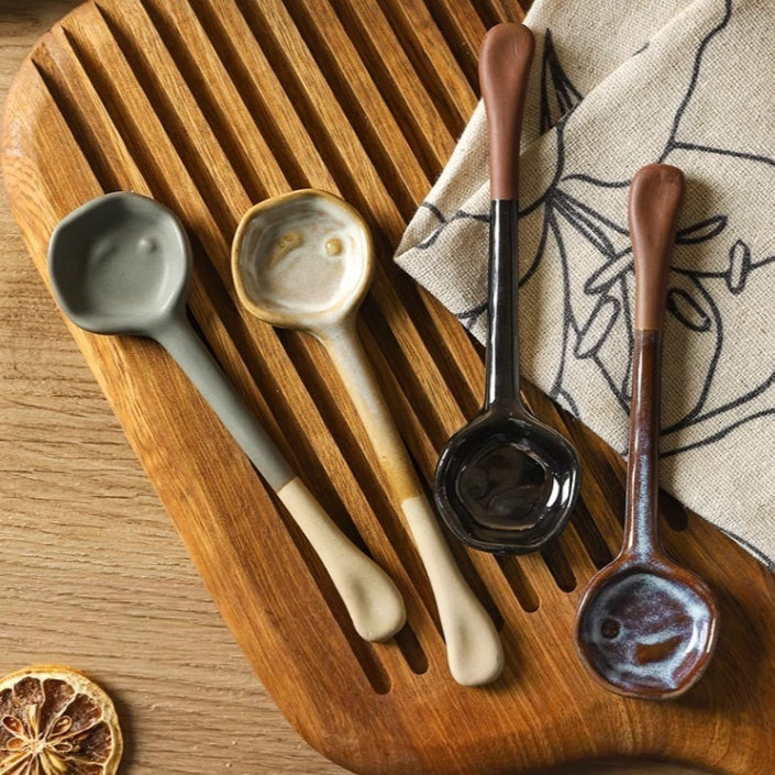 Four long-handle ceramic spoons in gray, beige, glossy black and blue-brown glazes on a slatted wood board
