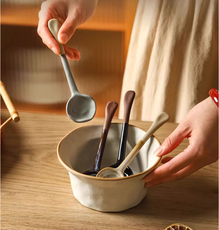 Hand lifting a gray ceramic spoon from an off-white bowl holding three more long-handle spoons, including a beige one