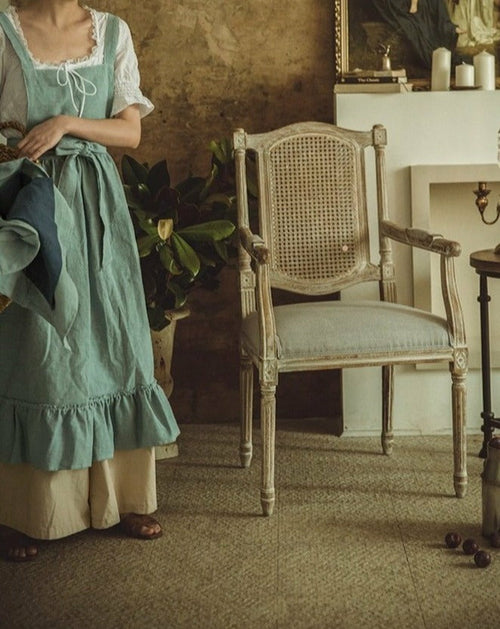 Sage green cotton-linen pinafore apron with a ruffled hem, worn over a long dress