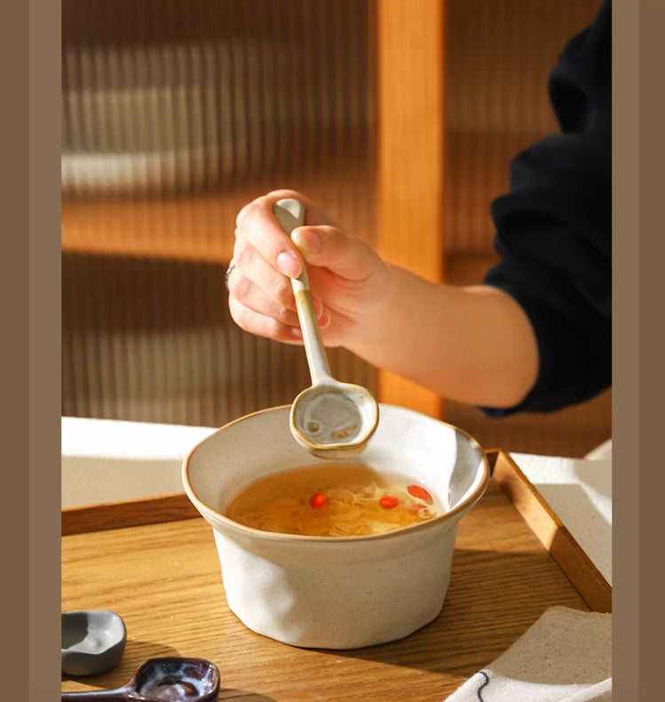 Hand using a beige ceramic long-handle spoon over a bowl of soup on a wood tray