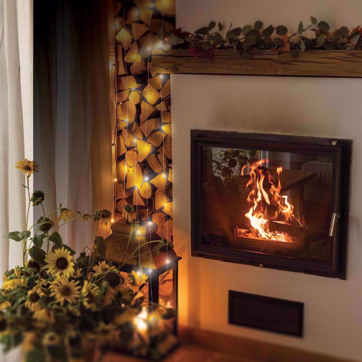 Warm-white fairy lights draped down a stacked firewood nook and into a lantern beside a lit fireplace